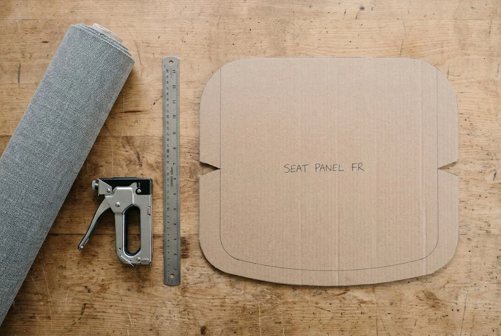 A cardboard seat panel template on a workshop bench with a fabric roll, a staple gun and a steel rule